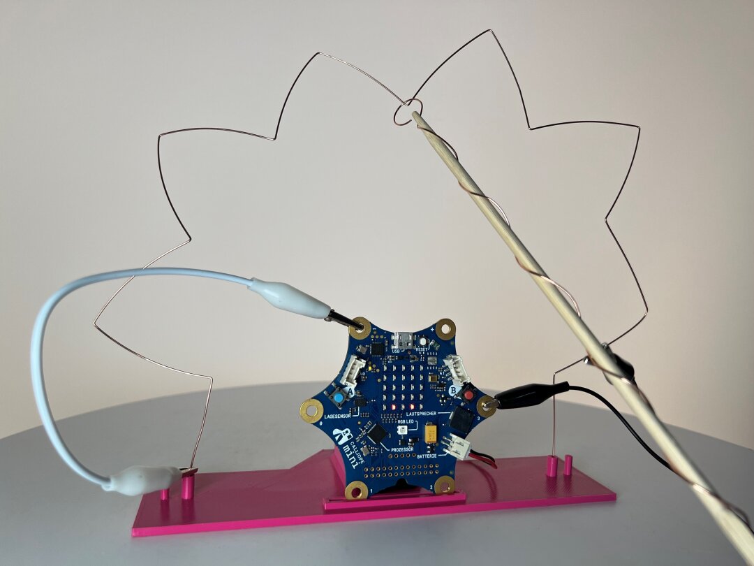 Calliope Mini - der heiße Draht Eine sternförmigen Platine steckt aufrecht in einer rosafarbenen Halterung. An ihr sind zwei Krokodilklemmen befestigt. Ein mit Draht umwickelter Holzstab hat eine Schlinge am vorderen Ende und wird um einen zackenförmig gebogenen Draht geführt.